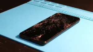 Smartfon z pękniętym ekranem leżący na niebieskiej macie serwisowej w profesjonalnym warsztacie.
